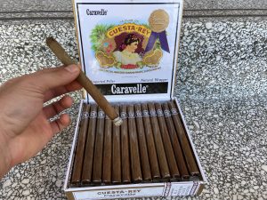 Open box of 25 count Cuesta Rey Caravelle cigars with hand holding lightly ashed one