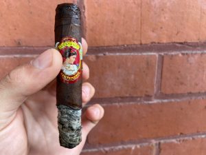 Hand holding Cuesta Rey No. 7 in front of brick wall with ash on the end